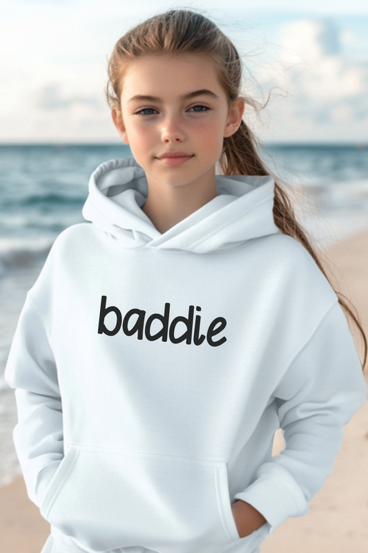 A young girl stands on a beach wearing a white hoodie with the word “baddie” in black lettering. The casual yet trendy style complements the relaxed seaside setting.