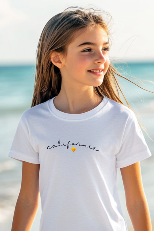 A young girl stands on a beach wearing a white “California” T-shirt with a small poppy design. This 100% organic cotton tee offers a breezy, stylish look for kids.