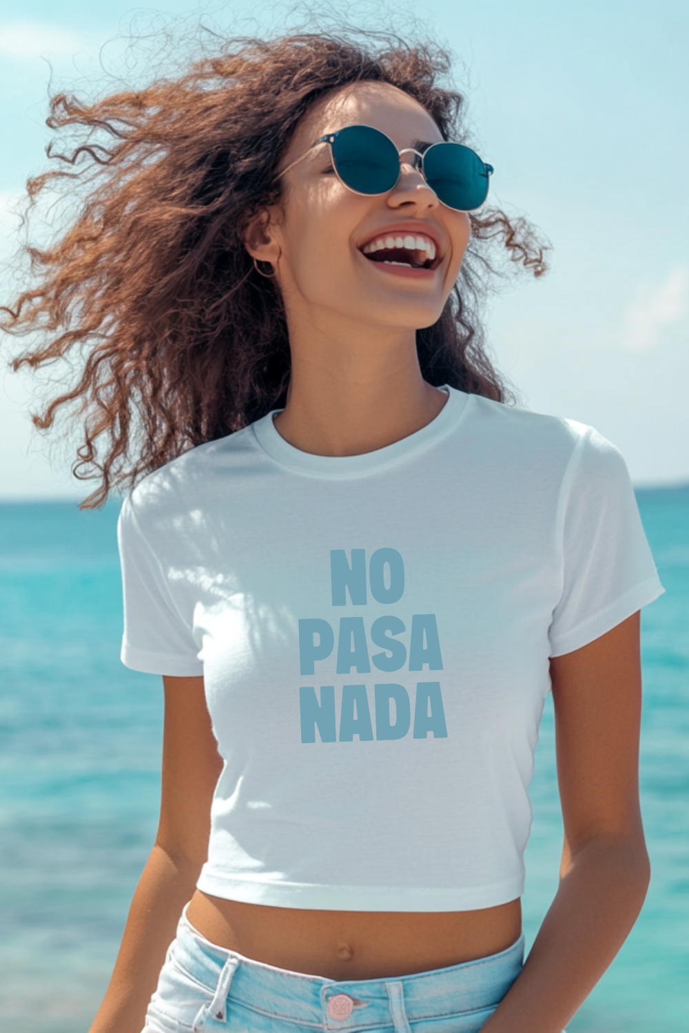 A smiling young woman wearing sunglasses stands by turquoise water, sporting a white baby tee that says “NO PASA NADA” in bold lettering. This unisex design works for both boys and girls, with a baby tee fit for women, offering a laid-back, Spanish-inspired style.