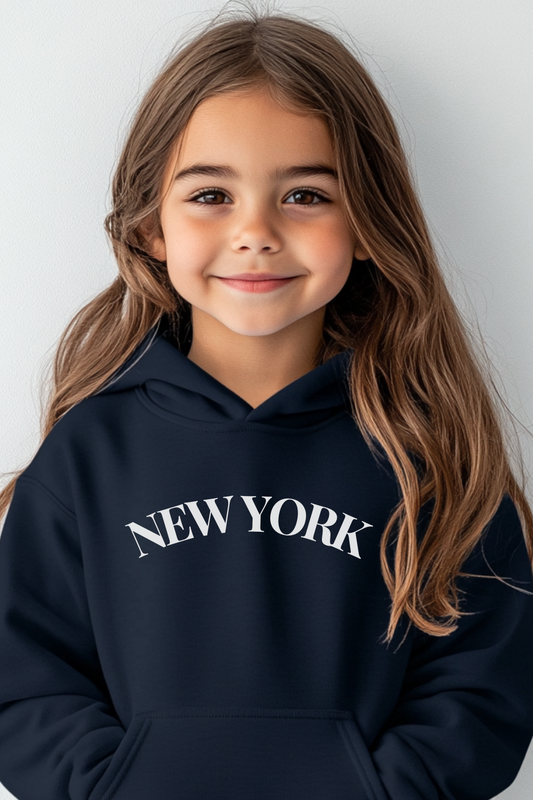 A young girl stands against a plain background wearing a navy hoodie with “NEW YORK” in white lettering, showcasing a trendy casual style.