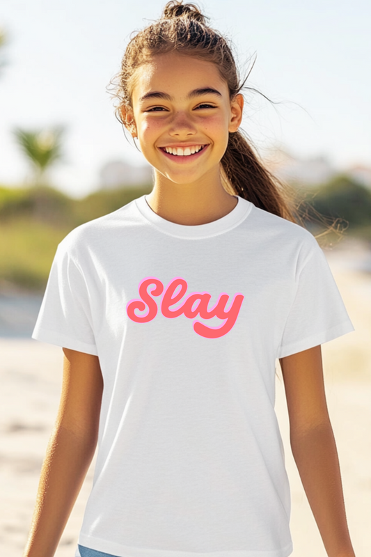 A smiling young girl stands on a sunny beach wearing a white T-shirt with a bold pink “Slay” graphic, perfect for trendy kids’ summer fashion.