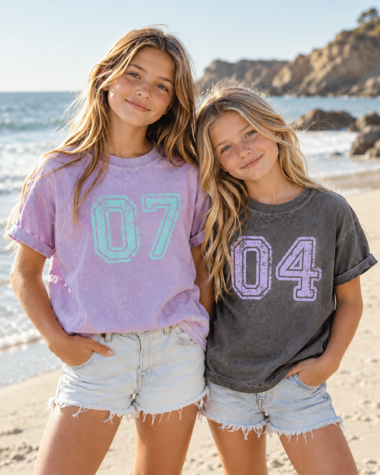 Two girls wearing AdaVie Threads custom varsity number T-shirts on a California beach. One wears a lavender Comfort Colors tee with an aqua 07, and the other wears a washed charcoal tee with a lavender 04.