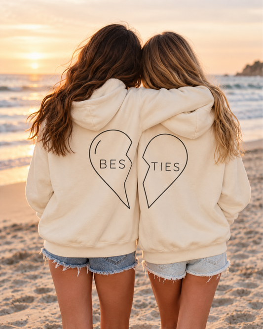Two best friends wearing matching ivory BES and TIES split-heart hoodies with their arms around each other on the beach at sunset.
