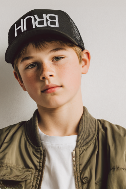 A tween boy wearing a black Yupoong retro trucker hat with bold upside-down “BRUH” white embroidery. Styled with a casual bomber jacket and white tee, the photo showcases modern streetwear for kids and teens. Perfect for product listings, youth fashion, or trendy meme-inspired accessories.