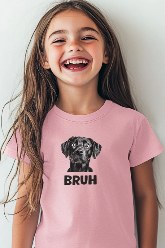 A young girl with long brown hair and a big smile is wearing a light pink t-shirt featuring a black-and-white graphic of a surprised-looking dog with wide eyes. Below the dog, the word "BRUH" is printed in bold black capital letters. The background is a simple, neutral-colored wall. The image conveys a playful and humorous vibe, making it ideal for youth fashion, funny pet-themed apparel, and expressive statement tees.